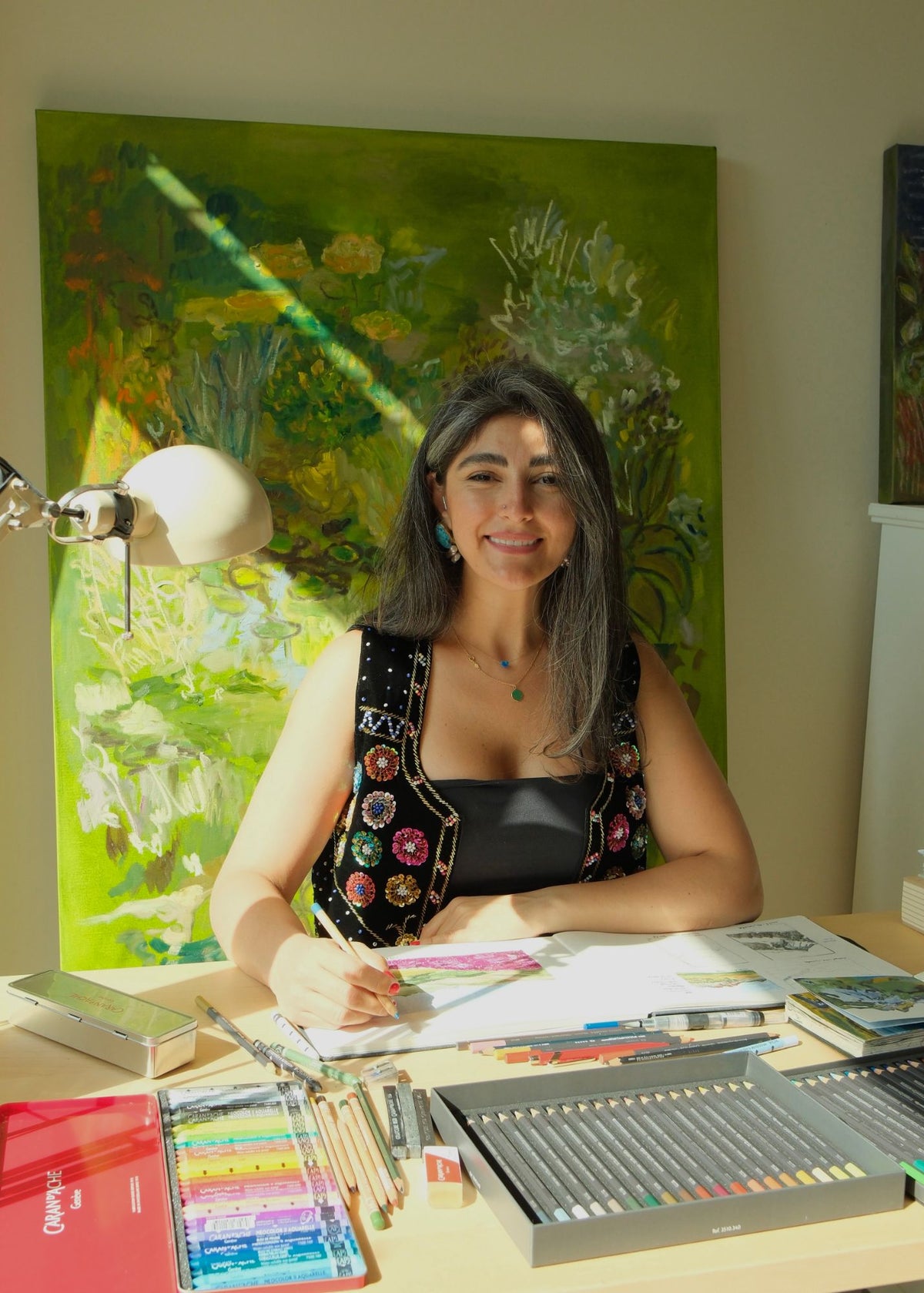 Fifi Pourmohseni drawing with coloured pencils at her desk, in front of one of her large green paintings
